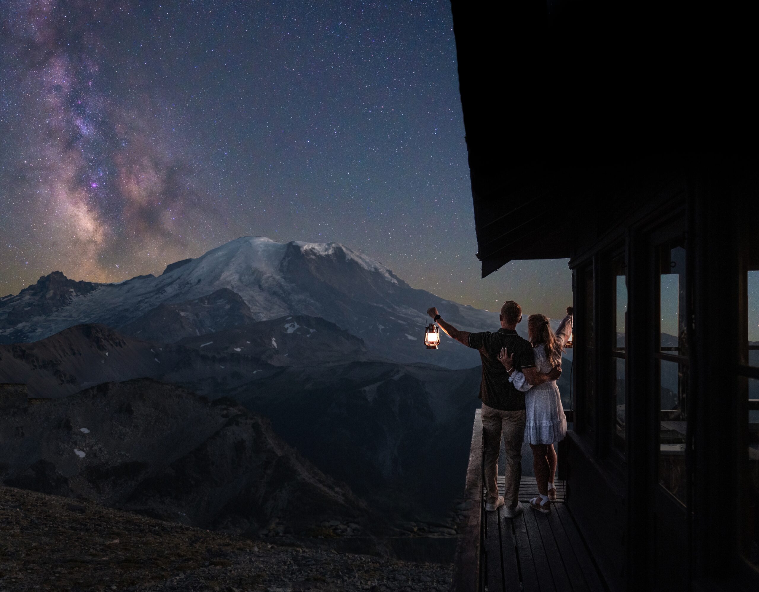 A couple standing on a fire lookout tower look out at the Milky Way rising over Mount Rainier